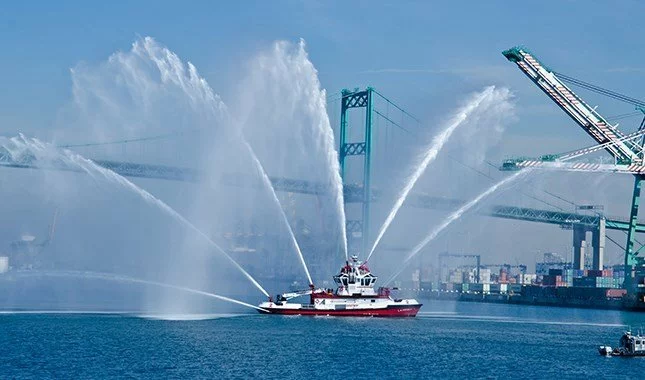 A fireboat sprays large streams of water into the air at a busy port, with cranes, shipping containers, and a bridge visible in the background under a clear blue sky.