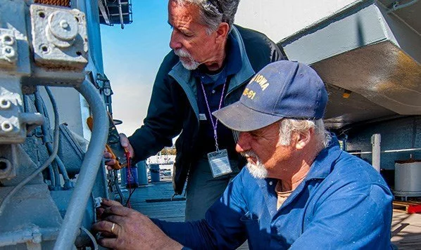 Two men work together on metal equipment outdoors. One man kneels, wearing a navy hat and blue shirt, while the other stands, holding a tool. Both focus closely on the machinery on what appears to be a ship.