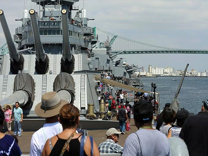People tour a large battleship docked at a harbor, with massive gun turrets visible. A bridge and industrial buildings are in the background under a partly cloudy sky.