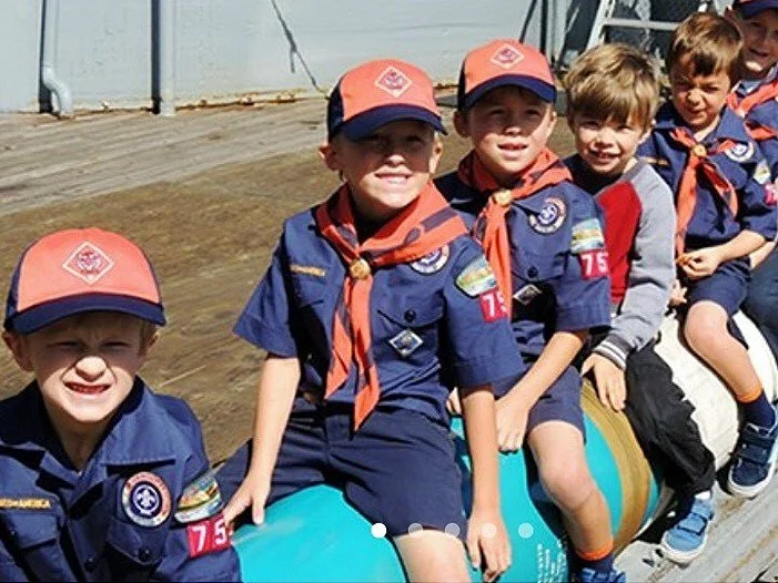 A group of young boys in blue Cub Scout uniforms and orange hats sit in a row outdoors, smiling at the camera. One boy in a gray jacket sits among them. The background shows a wooden deck and railing.