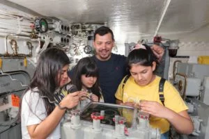 A man and three girls examine instruments inside a submarine or ship, surrounded by mechanical equipment and pipes. A man and three girls examine instruments inside a submarine or ship, surrounded by mechanical equipment and pipes.
