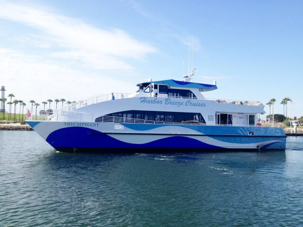 A blue and white Harbor Breeze Cruises boat named Triumphant floats on calm water with palm trees and a lighthouse visible in the background under a clear sky, offering a unique perspective of the vibrant waterfront near the Los Angeles Museum district.