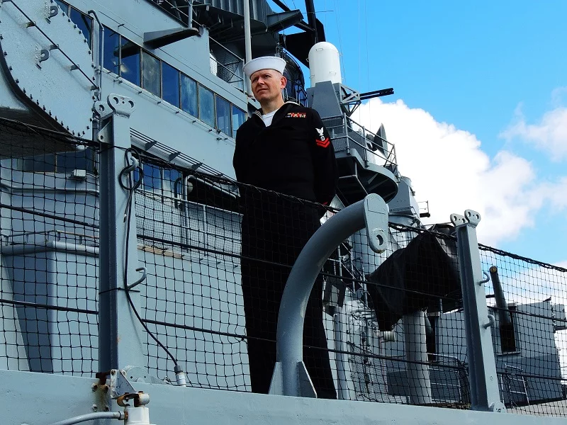A Navy sailor in uniform stands at attention on the deck of a gray military ship at the Los Angeles museum, with metal rails and a net fence in the foreground and blue sky in the background.