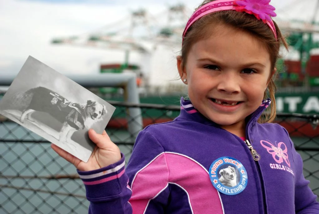 A smiling girl named Vicky in a purple jacket holds a black-and-white photo of a dog. She wears a button with the dog’s picture on it. Industrial structures and water are visible in the background.