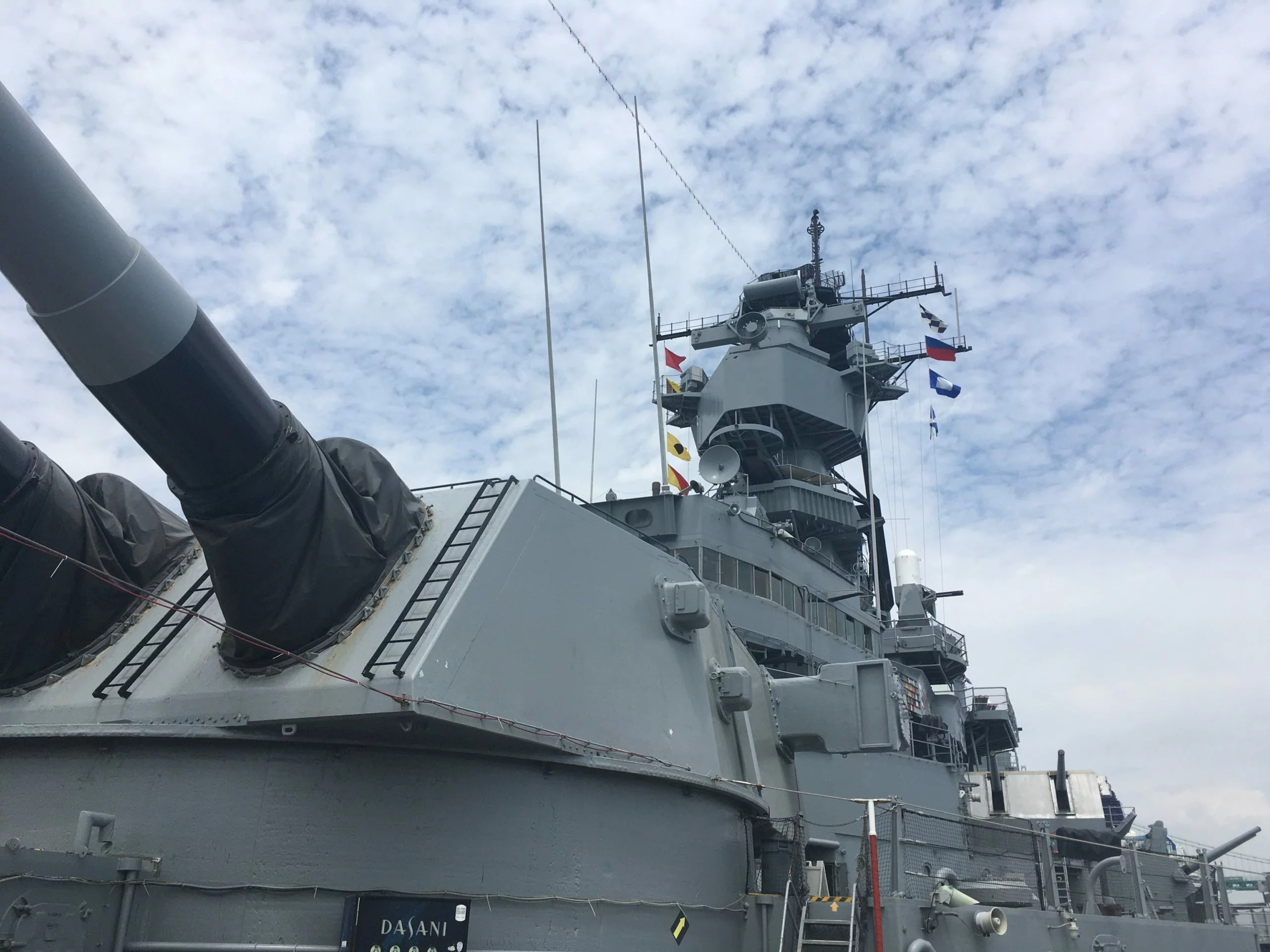 Close-up view of Battleship IOWA’s turret and superstructure under a partly cloudy sky, with several flags flying from the mast—capturing a striking moment in this latest Battleship IOWA Update.