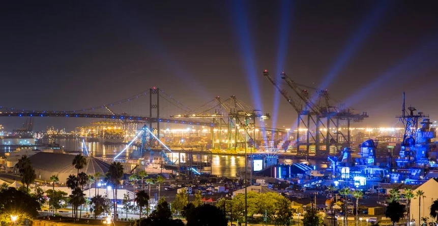 A brightly lit port at night with cranes, shipping containers, a bridge, and blue spotlights beaming into the sky. Ships and various buildings are visible along the waterfront, creating a bustling scene reminiscent of the coordinated energy seen during Operation Crush COVID.