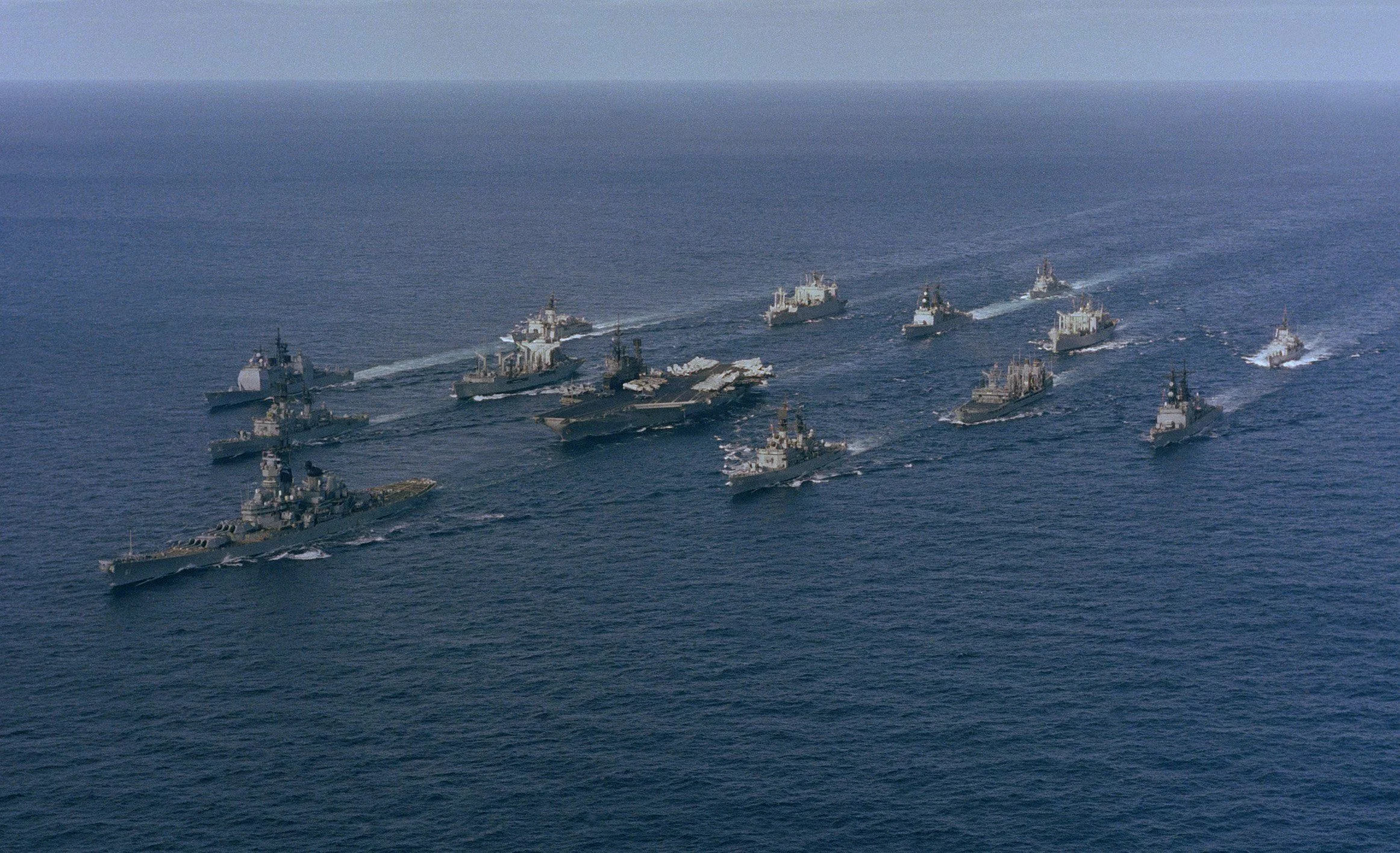 Surface Navy Aerial view of a naval fleet, including aircraft carriers and warships, sailing in formation across open water under a clear sky.