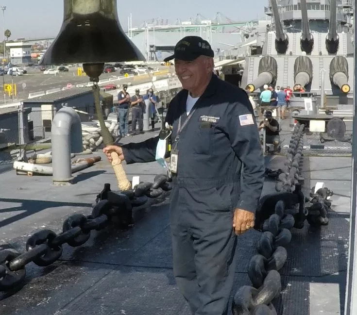 An older man in a navy blue jumpsuit and cap stands on a ship’s deck, smiling and holding a rope attached to a large bell. Thick anchor chains run along the deck, and other people—including fellow Battleship IOWA volunteers—are visible in the background.