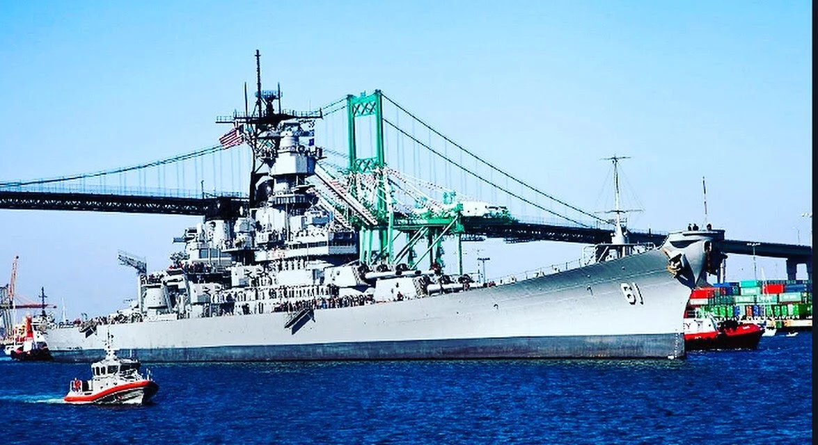 A large gray battleship is docked near a green suspension bridge, with smaller boats nearby and shipping containers visible in the background under a clear blue sky, offering a scene reminiscent of a CEO Update on bustling port operations and maritime strategy.