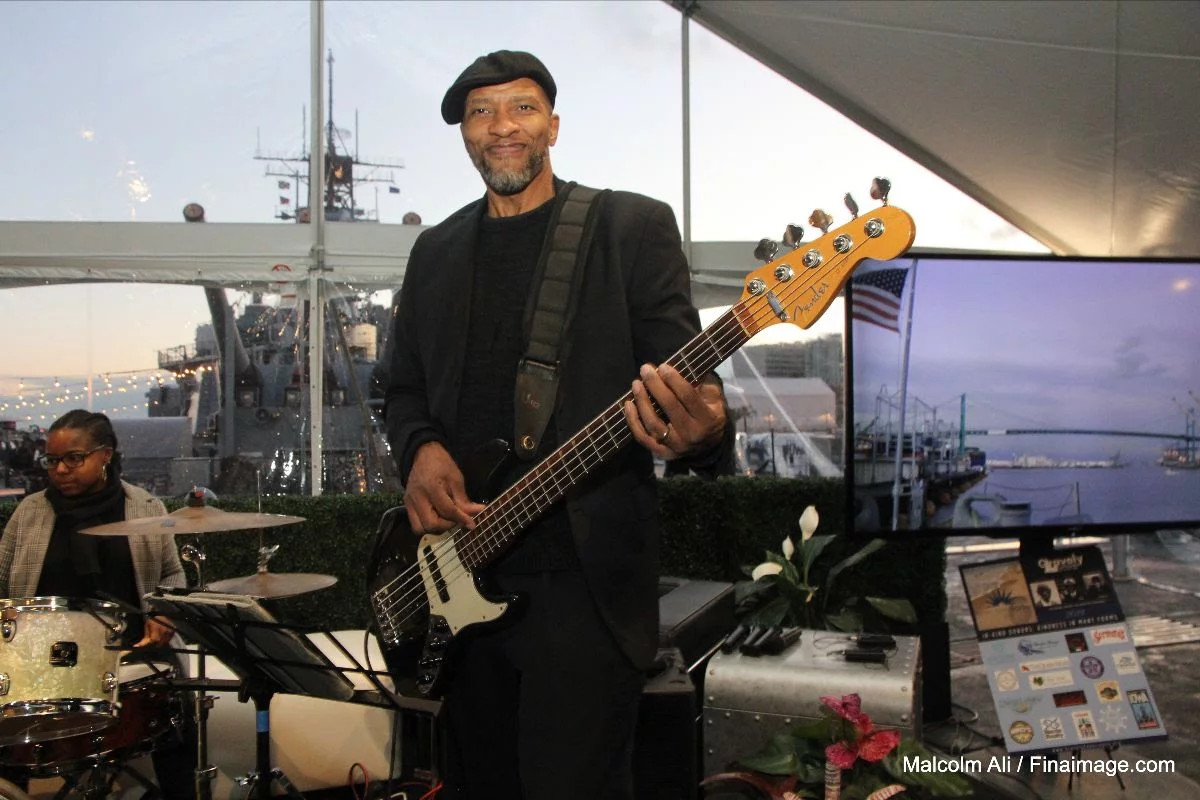 A smiling man wearing a black beret plays an electric bass guitar at an indoor Veterans Update event. A drummer sits nearby, and a ship with an American flag is visible through large windows behind them.