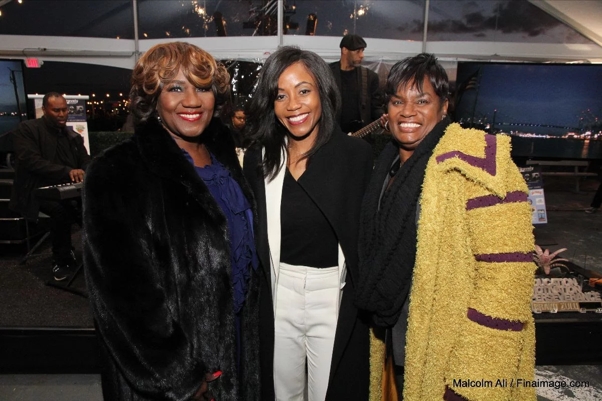 Three smiling women pose together indoors at a Veterans Update event. Two wear dark coats, and one wears a yellow and purple coat. A band performs in the background, while city lights twinkle through large windows behind them.