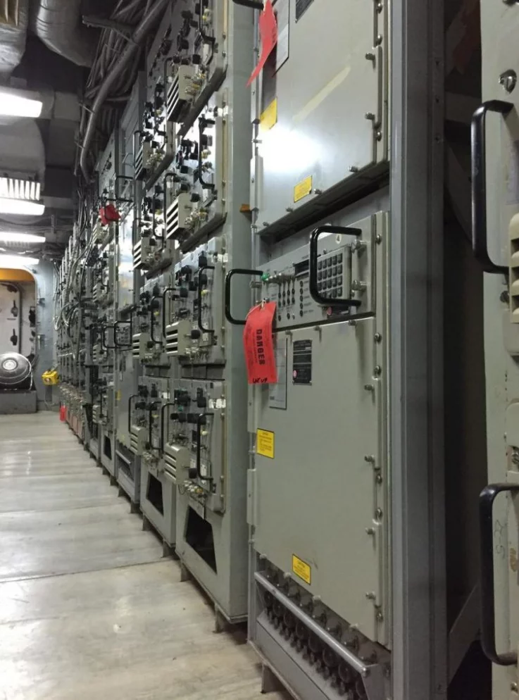 A row of large, gray industrial control panels with switches, dials, and warning labels inside a facility stands ready for operation. Announcements regarding system status are often posted nearby to ensure safety and awareness. One panel has a red tag attached to its handle. The floor and ceiling feature exposed industrial piping.