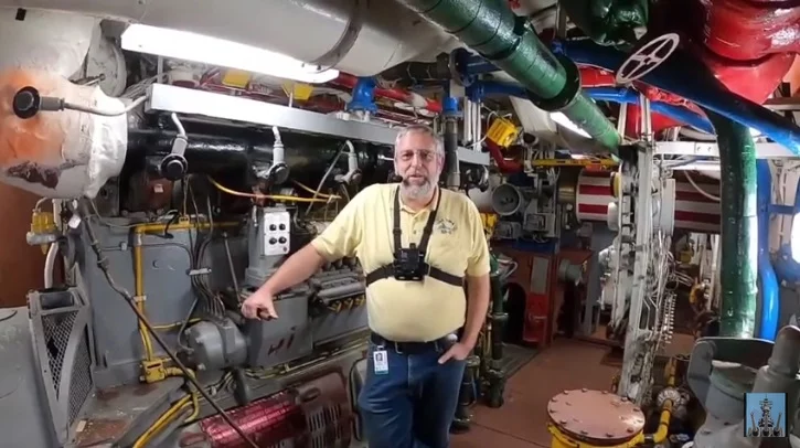 A man in a yellow shirt stands in a colorful, crowded engine room with machinery, pipes, and gauges. Wearing a camera harness and an ID badge, he looks toward the camera—capturing the environment as part of a special CEO Update.