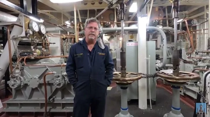 A man in blue coveralls stands in an industrial room with large pipes, gauges, and machinery. The area appears clean and organized, suggesting a professional work environment—an ideal setting for a CEO update on operational excellence and workplace standards.