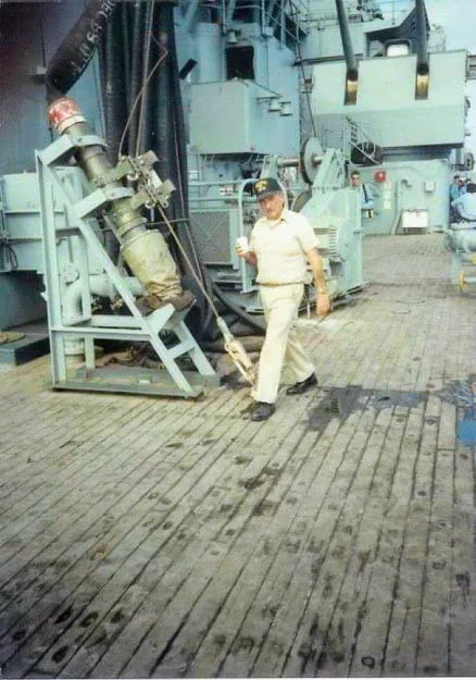 A person in a light uniform and cap walks on the deck of a naval ship, holding a drink, perhaps taking a moment to reflect after delivering the latest CEO Update. Large machinery and hoses are visible in the background.