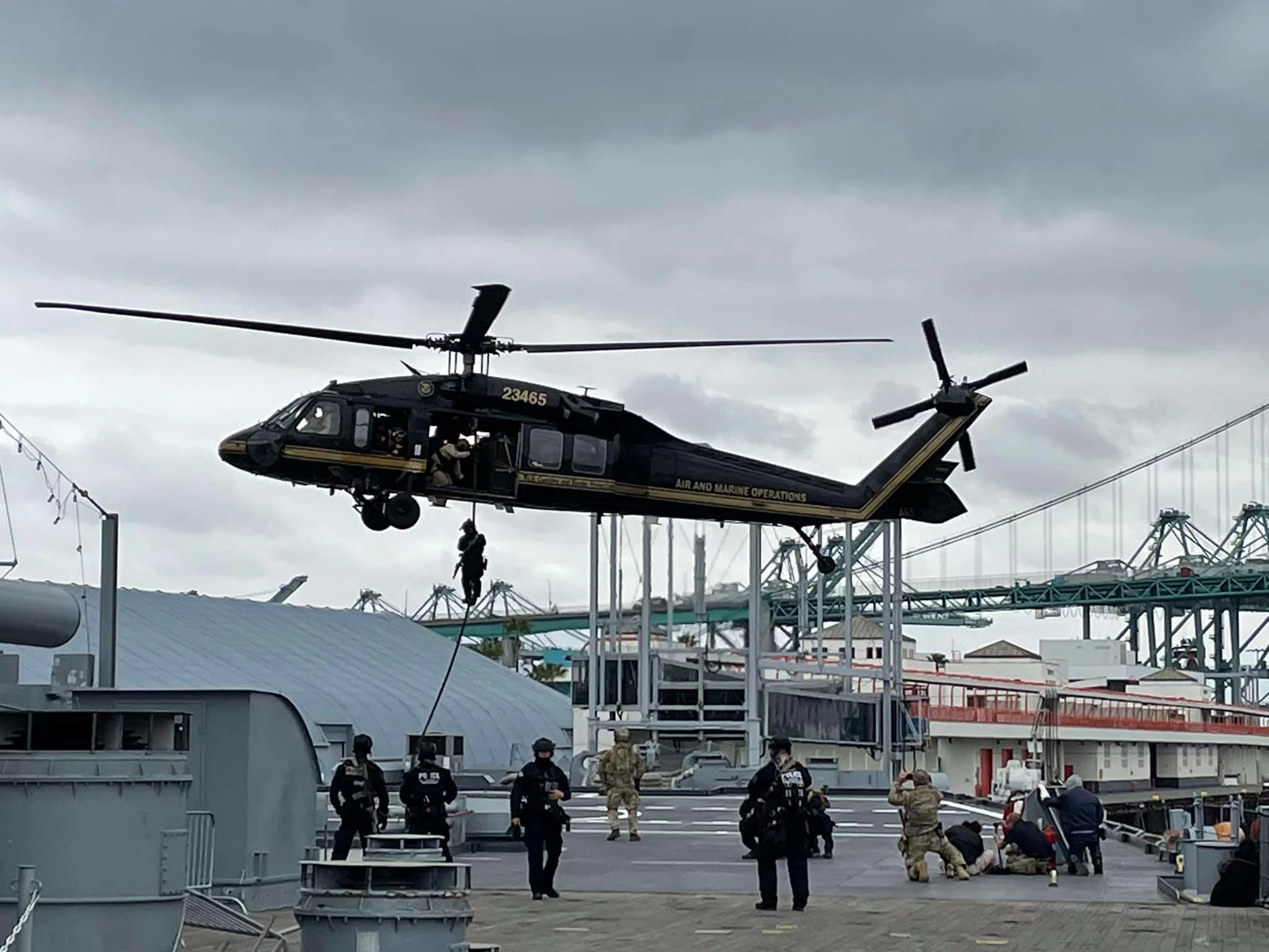 A black helicopter hovers above a ship as people rappel down ropes; several others in uniform stand and walk nearby. Overcast sky, bridge, and docked ships are visible in the background, setting a dramatic scene akin to critical moments highlighted in a CEO Update.