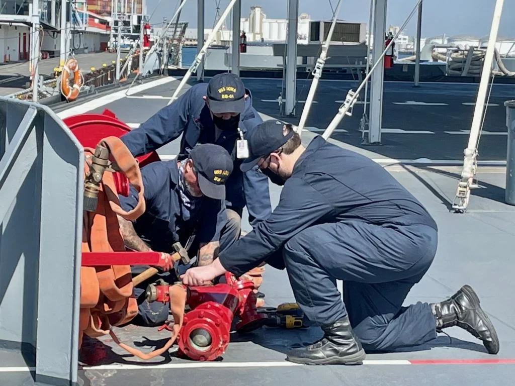 Three people in navy blue uniforms kneel and work on a red fire hose system on the deck of a ship, surrounded by pipes and safety equipment under a clear sky, as part of the Hull Preservation 2021 initiative. Three people in navy blue uniforms kneel and work on a red fire hose system on the deck of a ship, surrounded by pipes and safety equipment under a clear sky, as part of the Hull Preservation 2021 initiative.