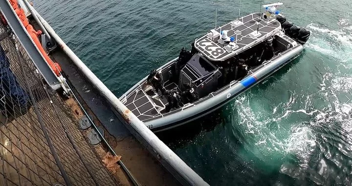 A black and gray patrol boat with the number 8221 is next to a dock, viewed from above. Several people in dark uniforms are on board, and the boat stirs the water as it maneuvers alongside the pier.