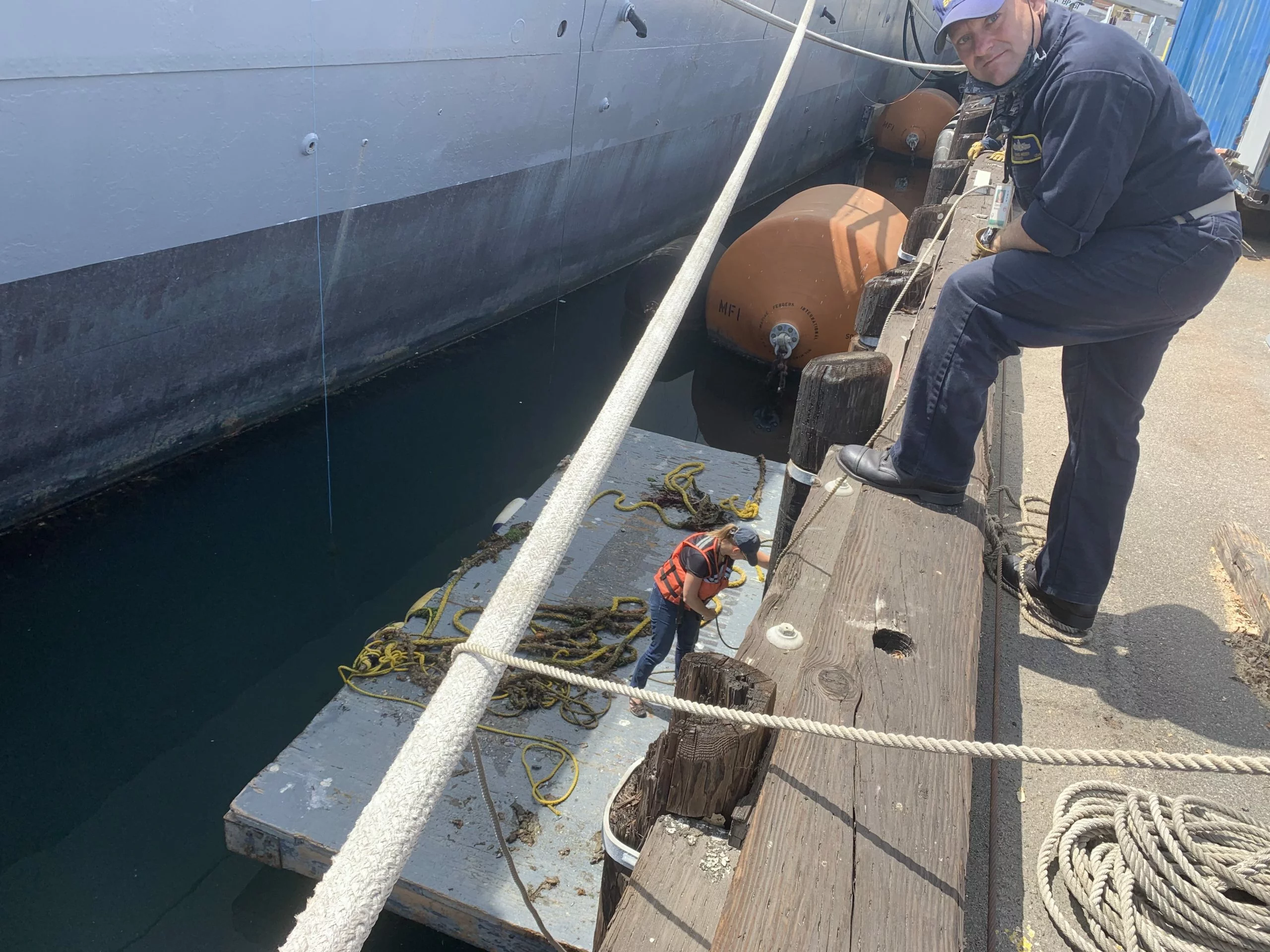 A person looks down from a dock, immersed in their own sea story, while another person in a life vest works with ropes on a floating platform next to a large ship.