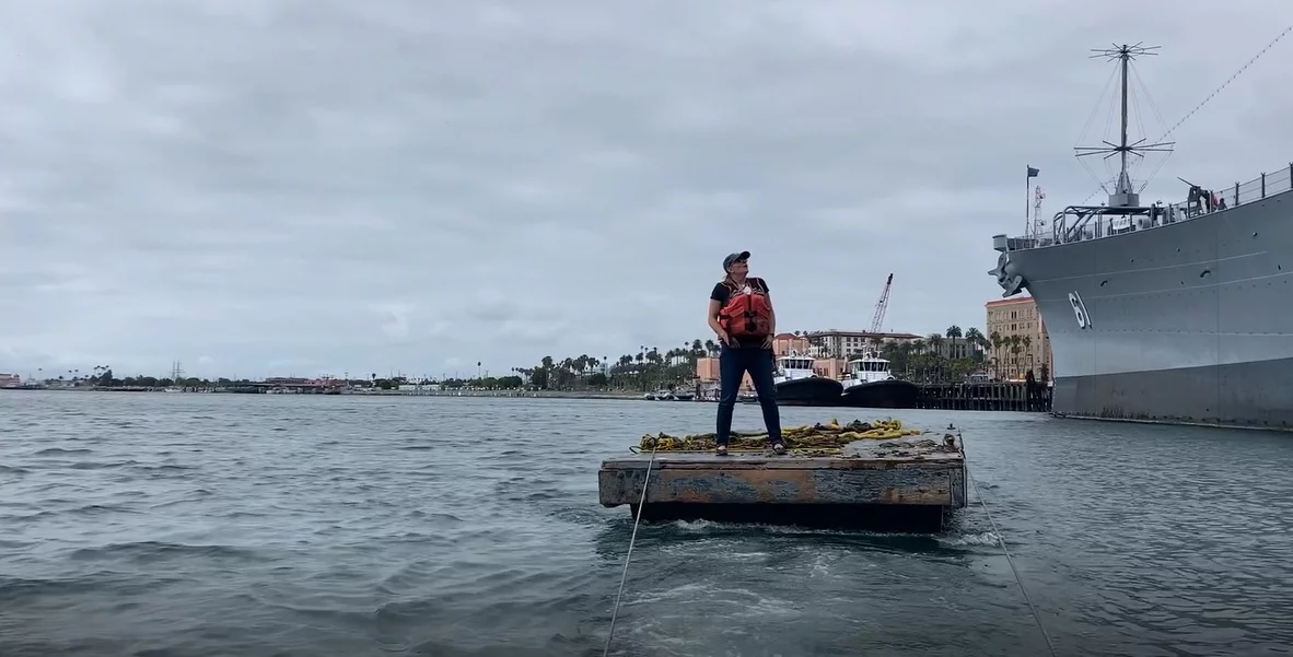 A person wearing a life jacket stands on a small, flat boat loaded with yellow items in a harbor, as if part of a real-life Sea Story, with a large gray ship and palm trees visible in the background under a cloudy sky.