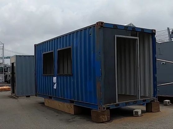 A blue shipping container with two windows and an open doorway sits on wooden blocks in an outdoor area, resembling the kind of workspace often highlighted in a CEO Update, with another container and industrial equipment visible in the background under a cloudy sky.