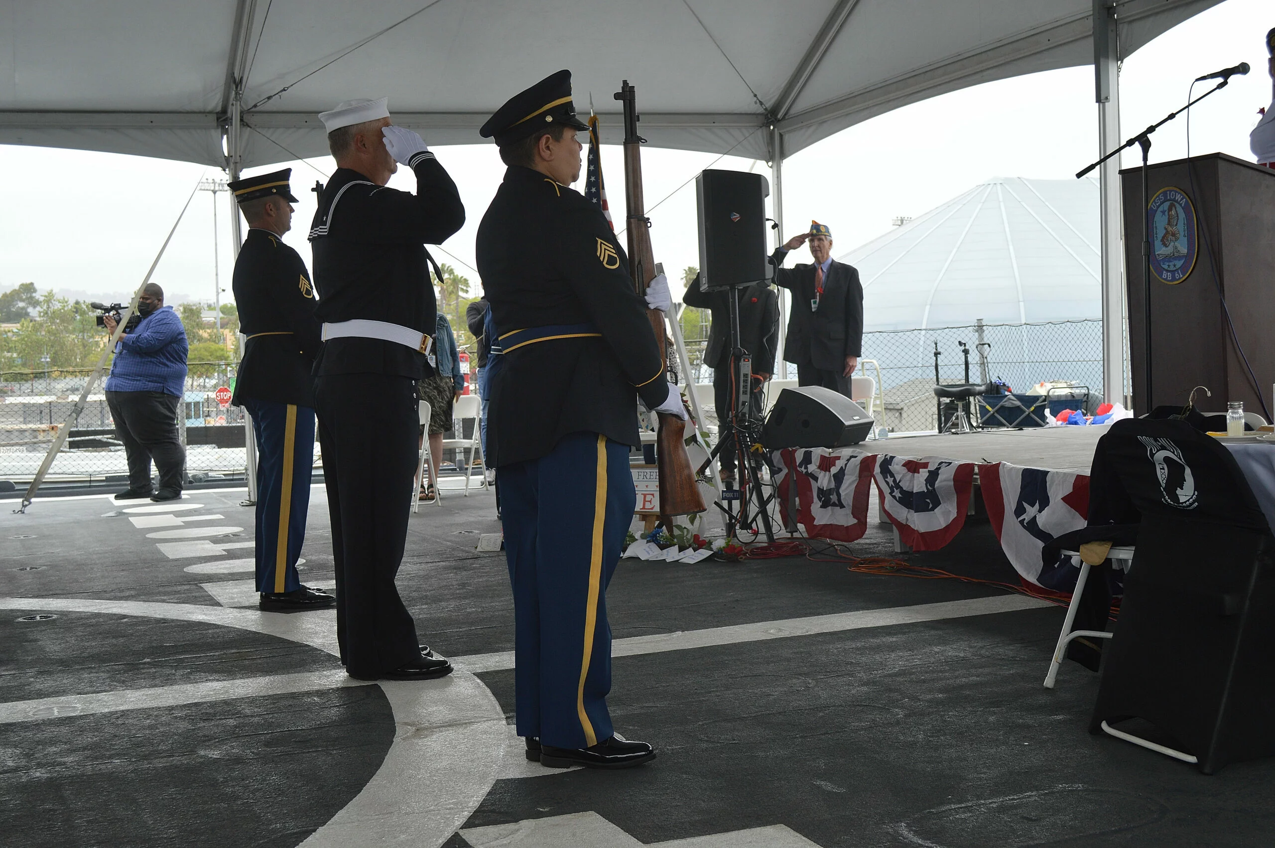 Three uniformed military personnel stand at attention and salute at a ceremony under a tent. A person stands at a podium nearby, delivering a CEO Update, with red, white, and blue bunting adorning the stage.