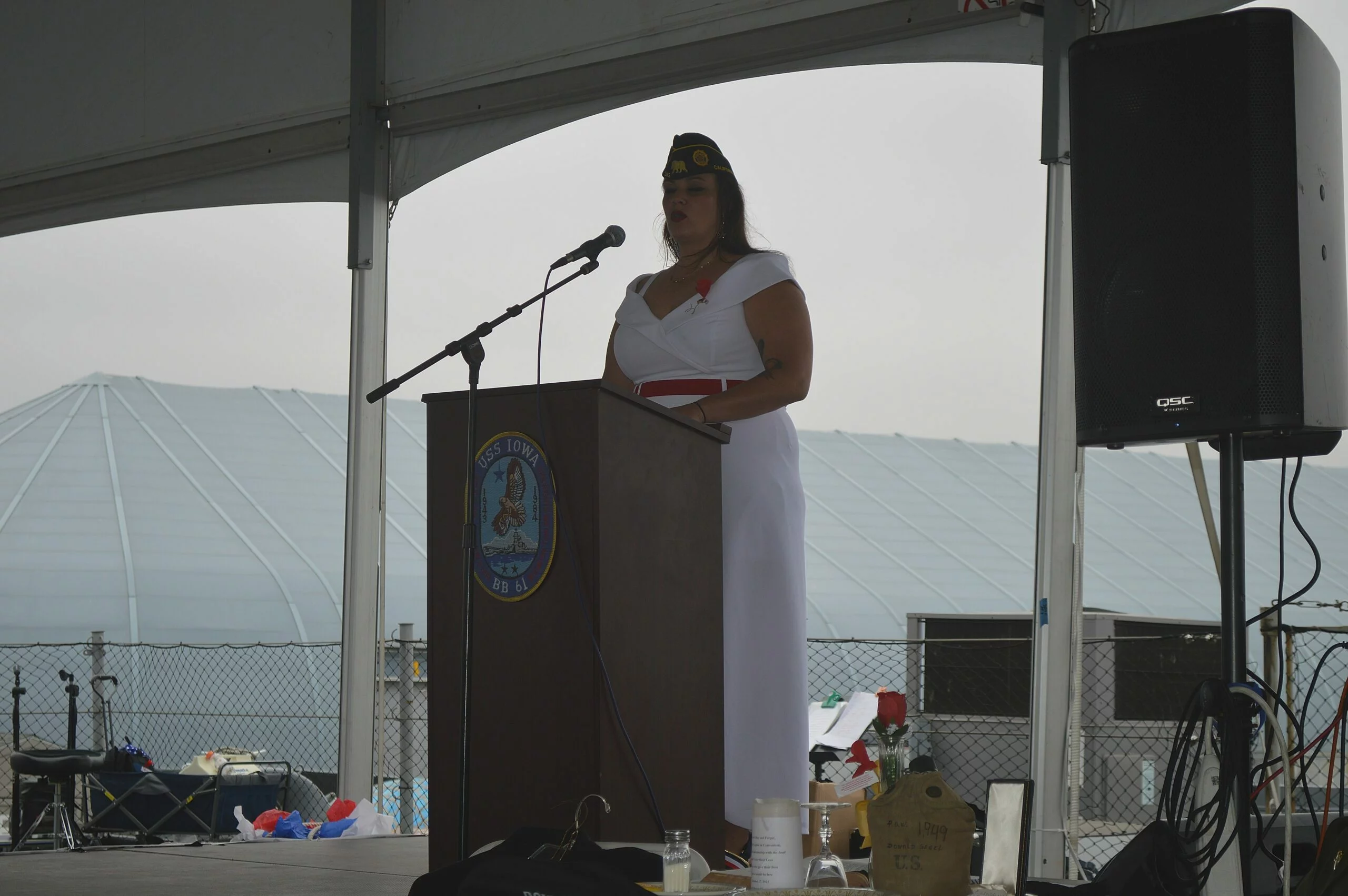 A woman in a white dress and military hat delivers a CEO Update at a podium on a covered outdoor stage. There are items arranged on the stage, with a large tent and fence visible in the background.