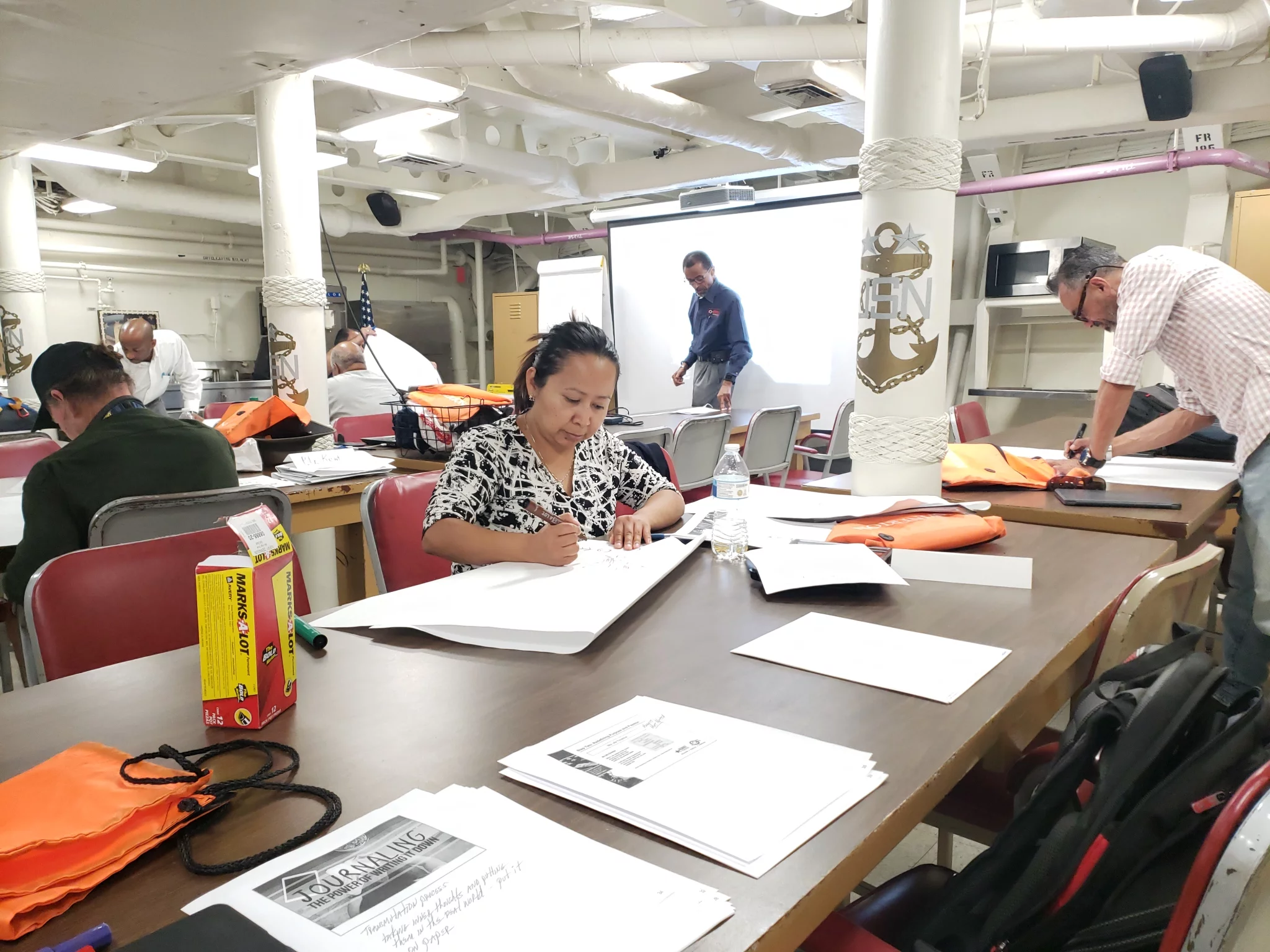 People, including several veterans, sit and work at tables in a classroom filled with papers and folders. One woman writes, while others stand or sit nearby. The room has fluorescent lights and a projector screen at the front.