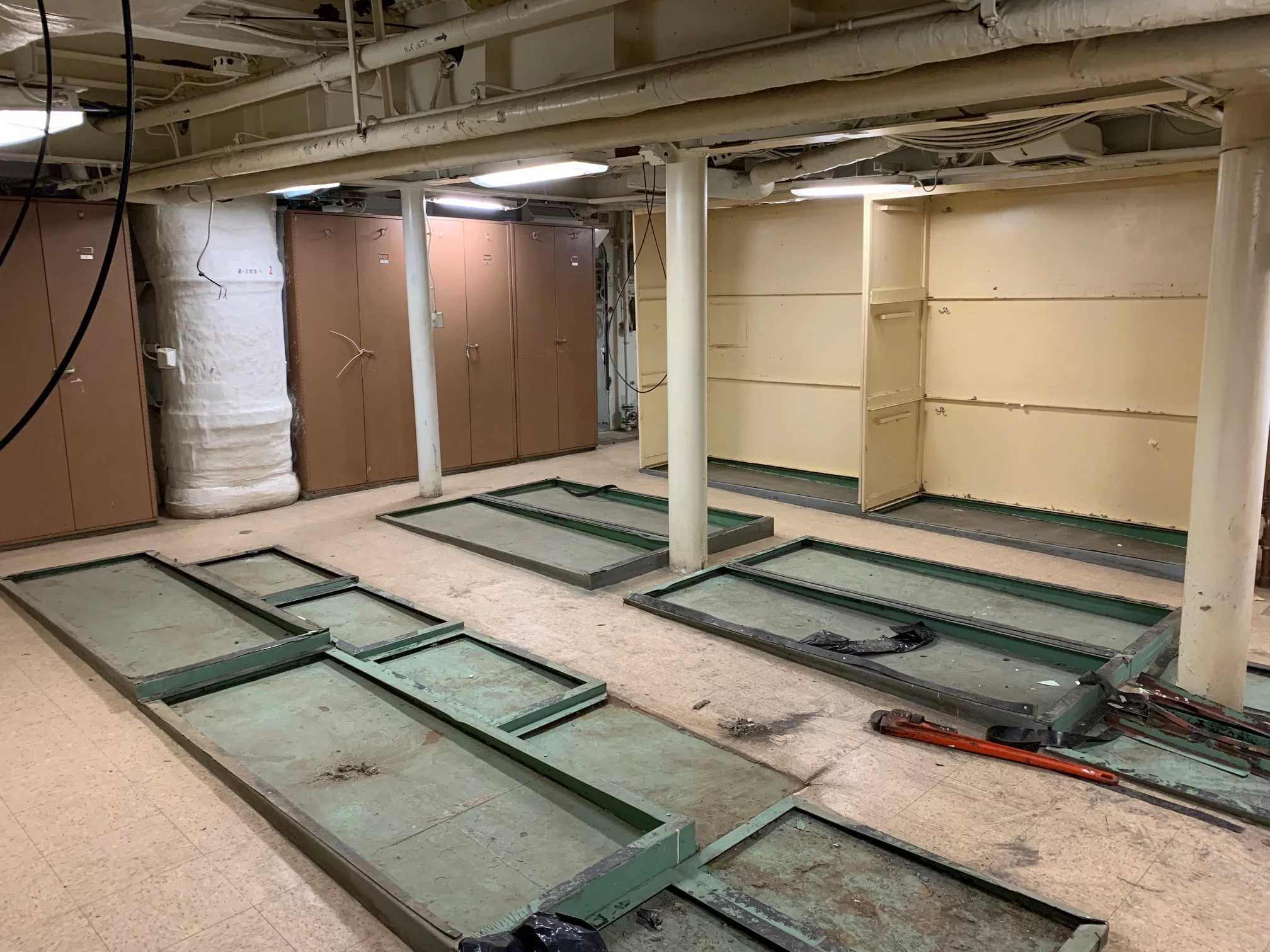 A room with exposed pipes, tan lockers, yellow walls, and green metal bed frames lying flat on the floor. The space appears to be old and somewhat industrial, reminiscent of a facility that might once have supported a Veterans Resource Center or offered veteran support services.
