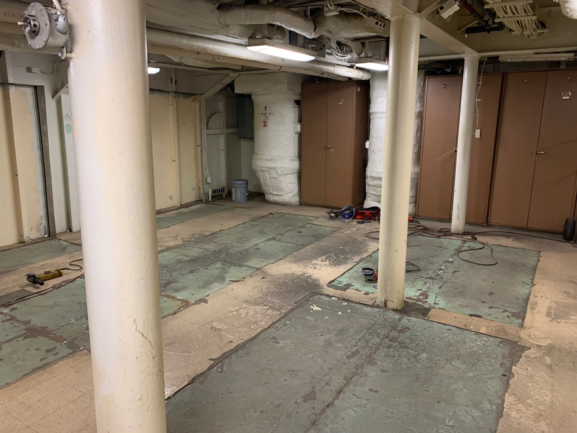 Industrial basement room with exposed pipes, support columns, peeling green floor tiles, brown storage cabinets, and scattered tools on the ground. The area appears to be under maintenance or renovation for a future Veterans Resource Center.