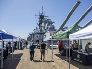 People, including veterans, walk between vendor tents set up on the deck of a large battleship, with cannons and ship structures visible in the background under a clear blue sky.