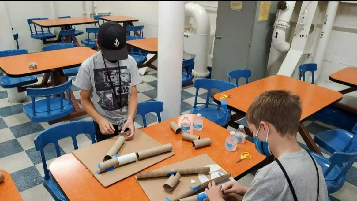 Two boys work on a craft project with cardboard tubes and tape at an orange table in a room with blue chairs. Water bottles, scissors, and cardboard pieces are also on the table.