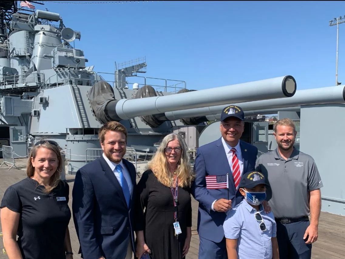 Six people, including a child holding an American flag, stand smiling in front of large naval guns on a battleship deck under a clear blue sky, proudly participating in the Welcome Sign Campaign.