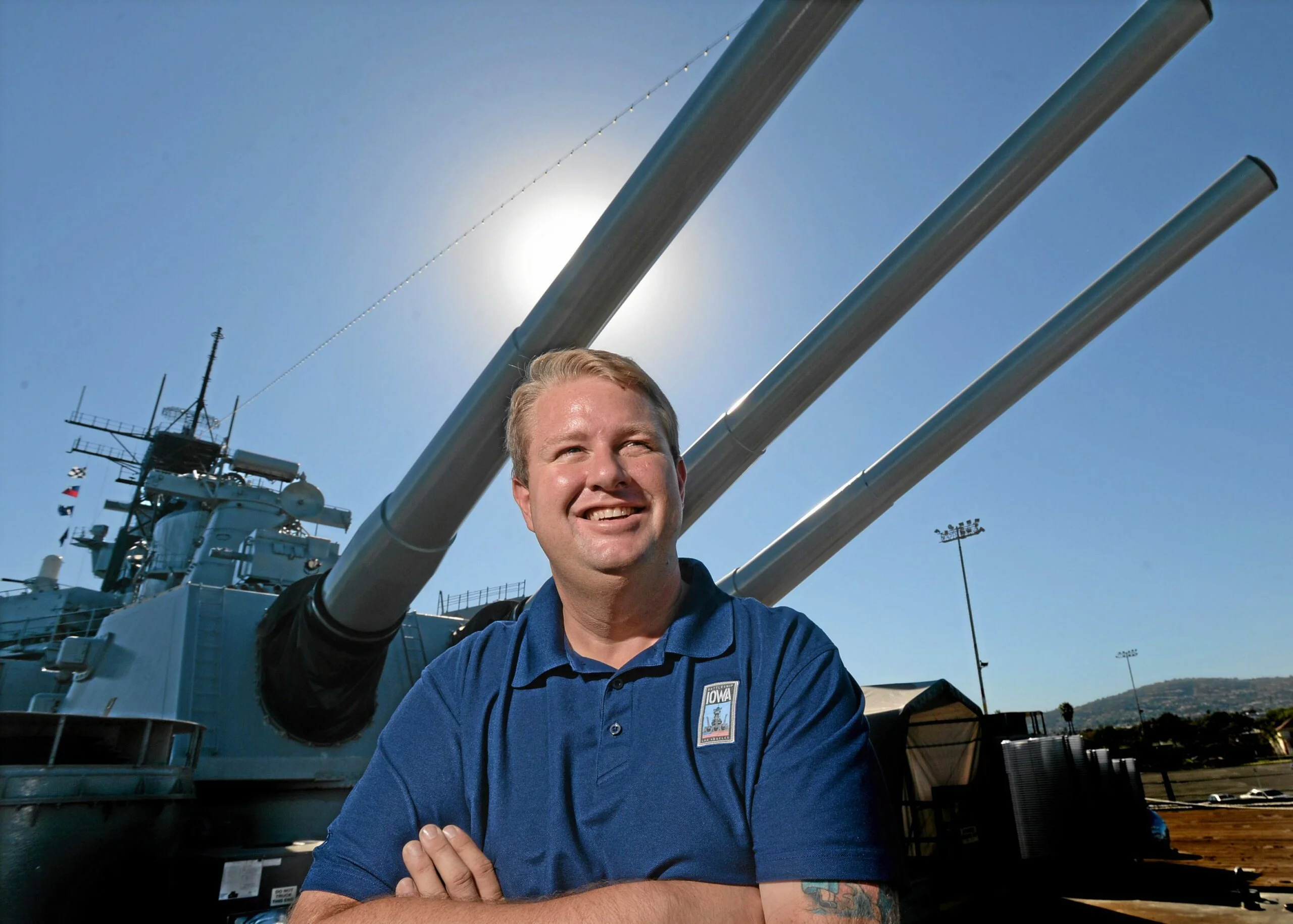 A smiling man in a blue shirt stands in front of a large battleship with three big cannons under a bright sky, the sun shining behind his head, as if posing for a CEO Update.
