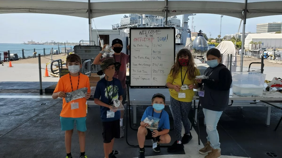 Six children wearing masks stand and kneel outdoors by a whiteboard under a tent, each holding objects wrapped in foil. A ship and ocean are visible in the background. The whiteboard welcomes participants to Battle Trip activities.