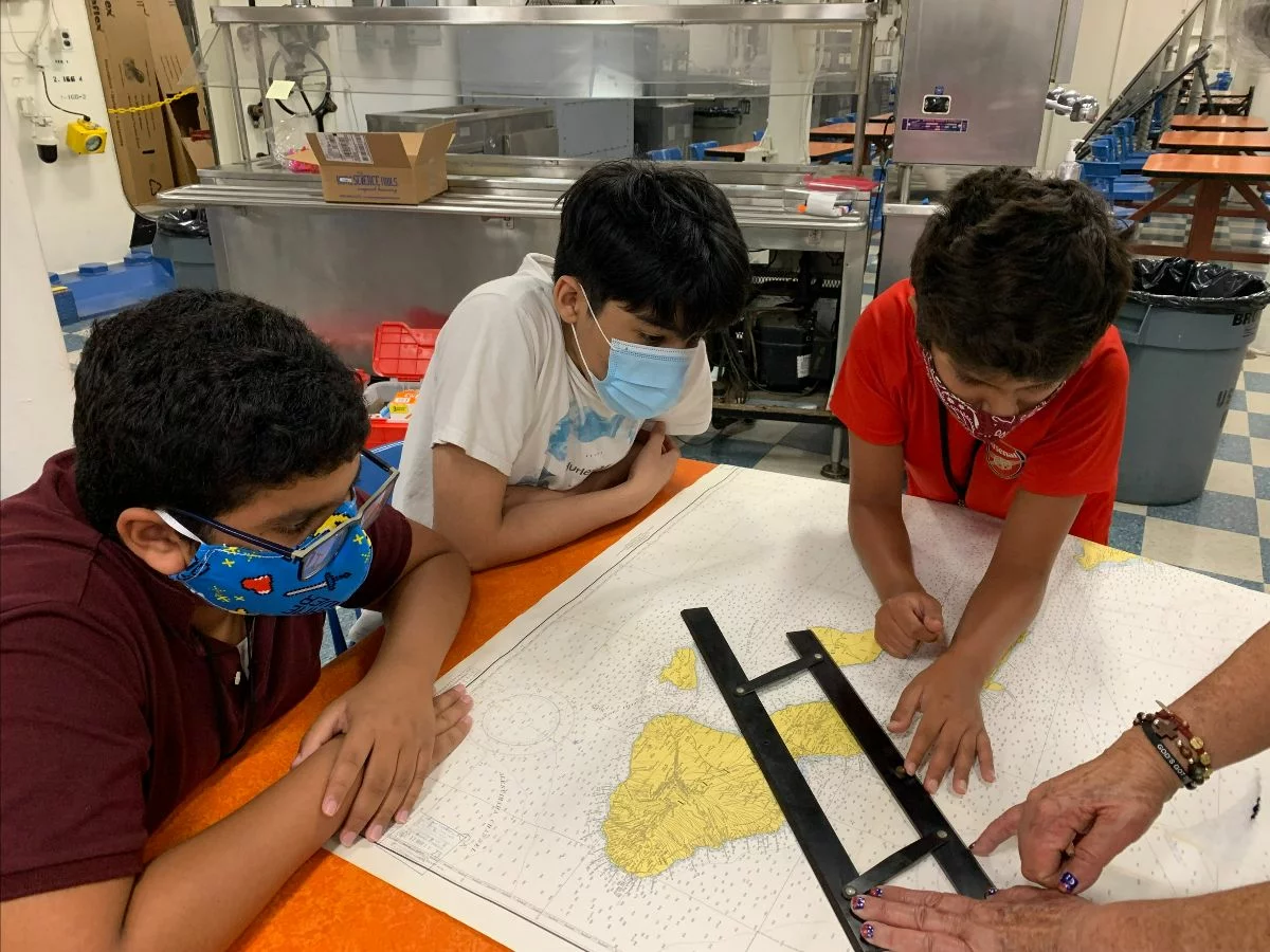 Three boys wearing masks look at a large map on a table while an adults hands use a ruler to measure. The scene appears to be in a classroom or lab with various equipment in the background.