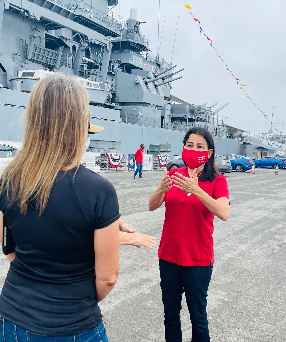 Two women talk near a large gray battleship. One faces the camera and gestures, wearing a red mask and shirt. The other woman, in a black top, faces away. Flags hang along the ship, and cars are parked nearby.