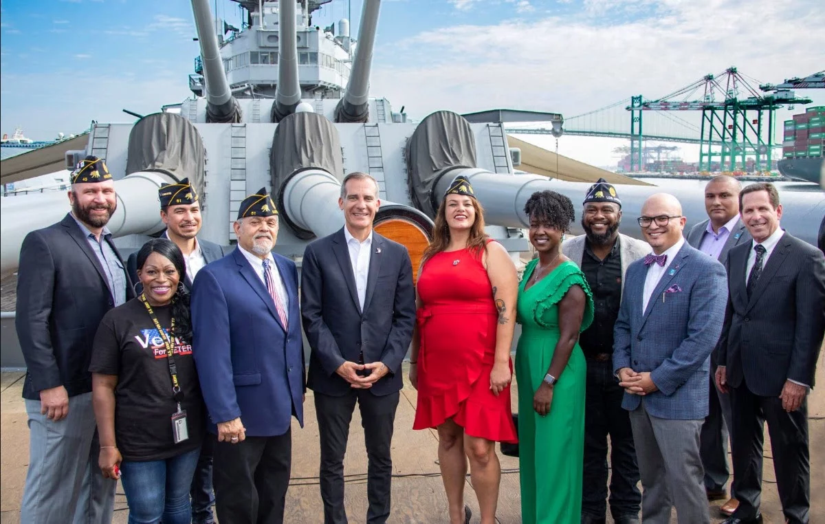 A diverse group of people, some in military hats, pose and smile together on the deck of a battleship with large cannons and machinery visible in the background under a partly cloudy sky.