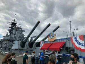 People gather near Vicky’s Doghouse, a food stand decorated with red, white, and blue bunting, in front of a large battleship with three long cannons, under a cloudy sky. People gather near Vicky’s Doghouse, a food stand decorated with red, white, and blue bunting, in front of a large battleship with three long cannons, under a cloudy sky.