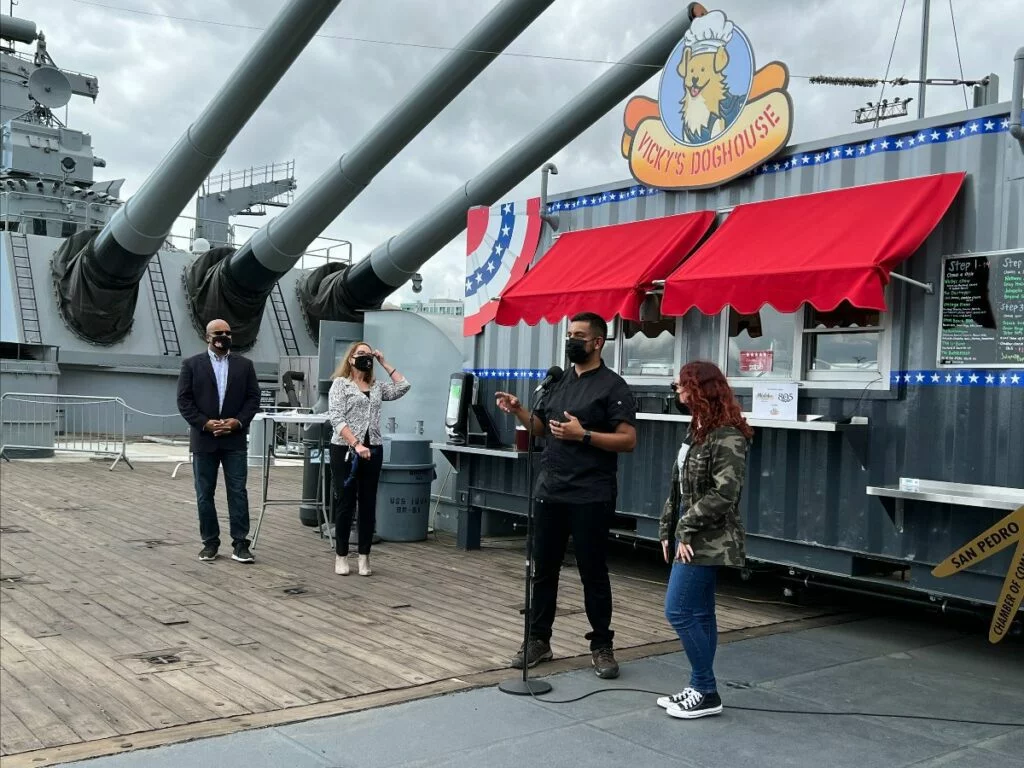 Four people stand on a ships deck near a food stand called Vickys Doghouse. Two large cannons are in the background. One person speaks into a microphone while others observe. Four people stand on a ships deck near a food stand called Vickys Doghouse. Two large cannons are in the background. One person speaks into a microphone while others observe.