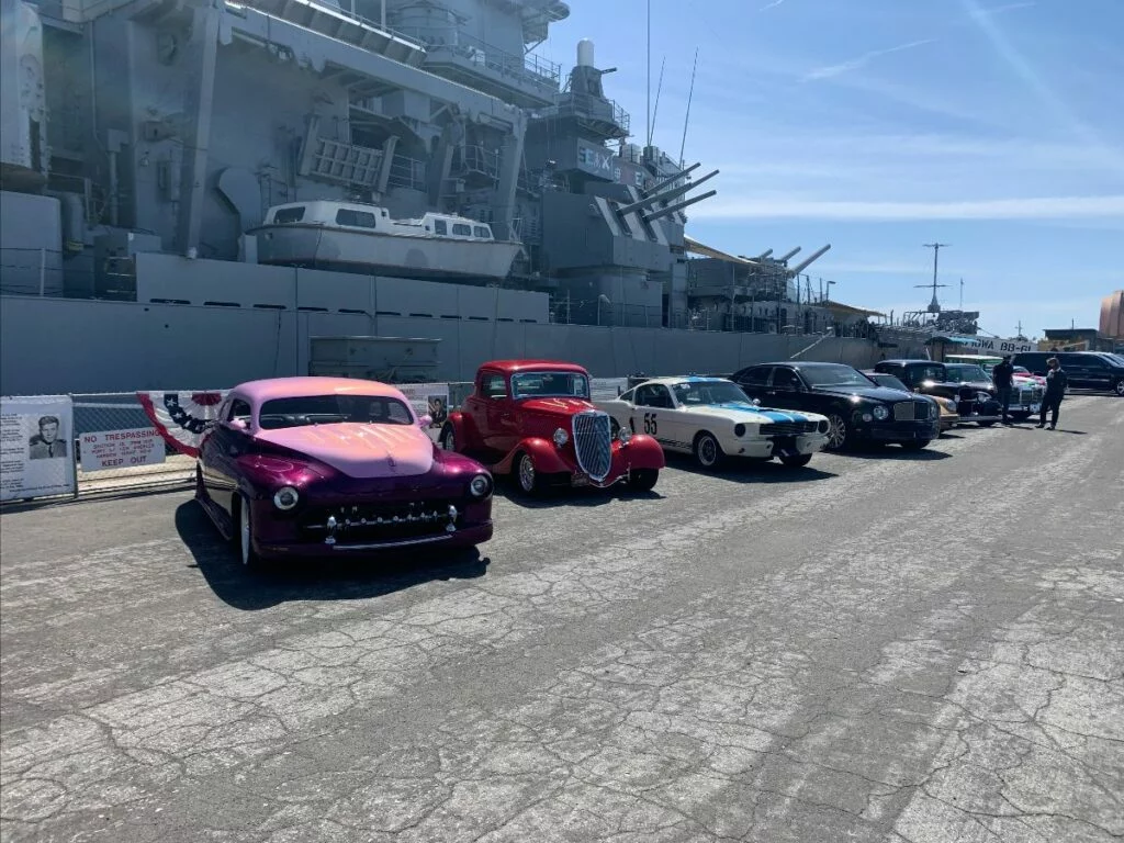 Five vintage cars are parked in a row on a cracked pavement near a large navy battleship, which is docked in the background. A few people are walking nearby under a clear, sunny sky. Five vintage cars are parked in a row on a cracked pavement near a large navy battleship, which is docked in the background. A few people are walking nearby under a clear, sunny sky.