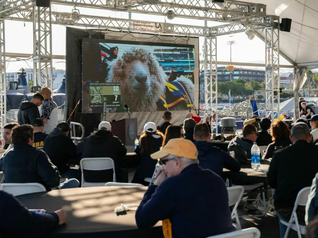 A group of people sit at round tables under a canopy, watching a large screen showing a close-up of a sheep at a sports event, with a scoreboard visible in the background—a unique scene featured in the latest museum update. A group of people sit at round tables under a canopy, watching a large screen showing a close-up of a sheep at a sports event, with a scoreboard visible in the background—a unique scene featured in the latest museum update.