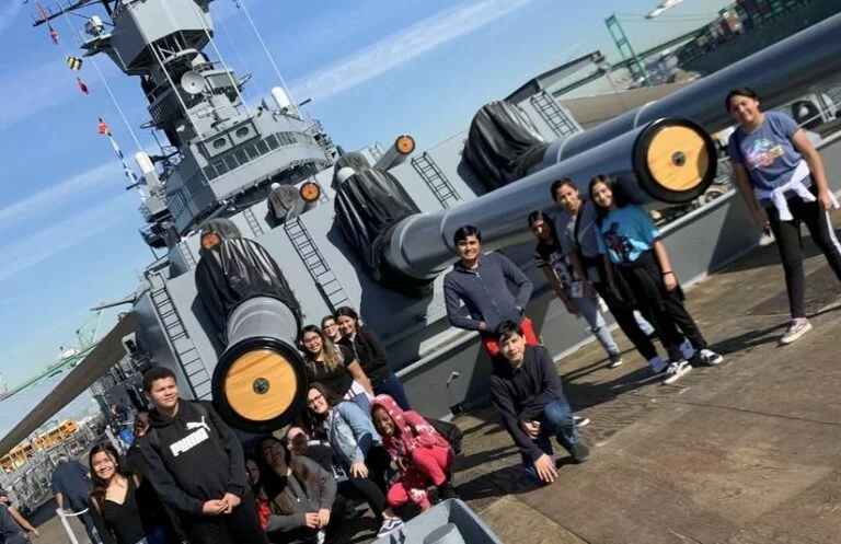 A group of teens and kids pose in front of a large gray battleship with big gun turrets on a sunny day in May 2023. Some are standing, some crouching, and everyone looks cheerful.