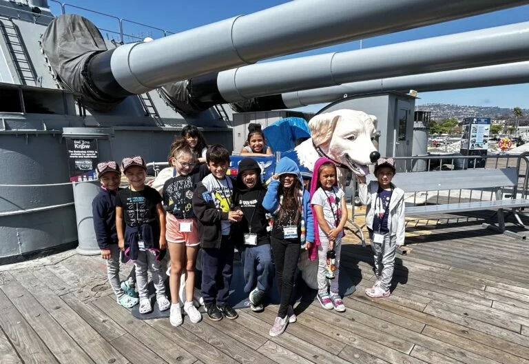 A group of children poses on a ship’s deck in front of a large white dog statue, with big gray cannons and city buildings visible in the background under a sunny blue sky—captured during the Museum Update on July 1, 2023.