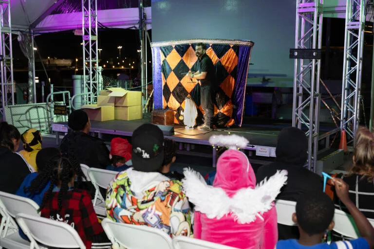 A performer stands on a stage with a checkered backdrop, entertaining an audience of seated children at events, some in colorful costumes, including an angel costume with wings and a hoodie with cartoon graphics.