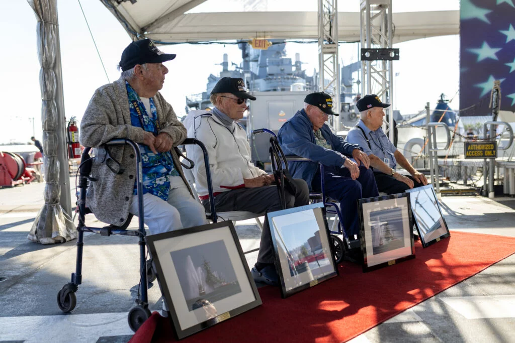Four elderly men wearing military caps sit in front of framed photos on a red carpet at an outdoor event near a battleship, under a tent with sunlight streaming in, as part of a Veterans Day commemoration. Four elderly men wearing military caps sit in front of framed photos on a red carpet at an outdoor event near a battleship, under a tent with sunlight streaming in, as part of a Veterans Day commemoration.