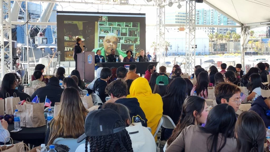 A large group watches a speaker and panel onstage under a tent, with a video call shown on a big screen behind them during the 2023 Museum Update. People sit at tables with paper bags, and city buildings are visible in the background.