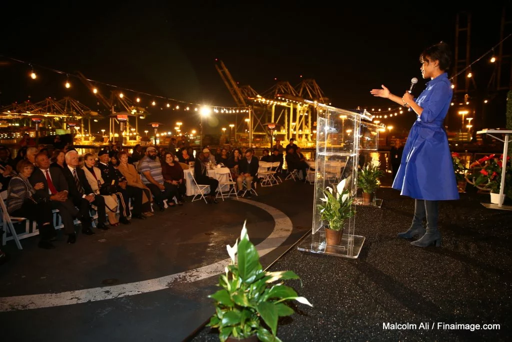 A woman speaks on an outdoor stage at night to an audience seated in front of her, with string lights overhead and industrial cranes—and the silhouette of a historic Battleship—visible in the background, setting the perfect atmosphere for an inspiring evening of education.