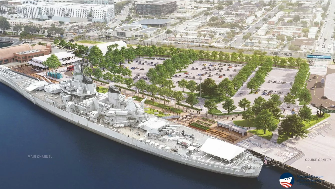 An updated scene shows a large battleship docked along a waterfront with nearby parking lots, trees, and city buildings in the background, viewed from above during daylight.