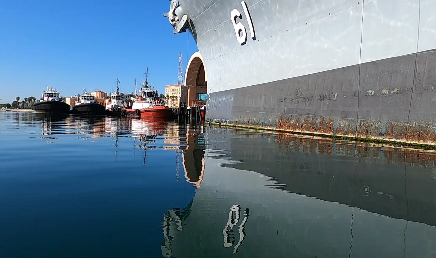 A large gray battleship marked 61, known as the IOWA, is docked in calm water, reflecting its massive hull. Smaller boats are moored nearby under a clear blue sky, with buildings visible in the background. Visitors are invited to donate and support the preservation of this historic vessel.